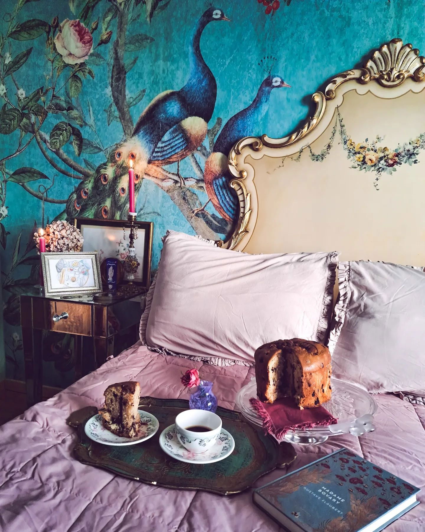 Cozy bedroom scene with cake, coffee, and "Madame Bovary" book on a pink bedspread. Peacock wallpaper.