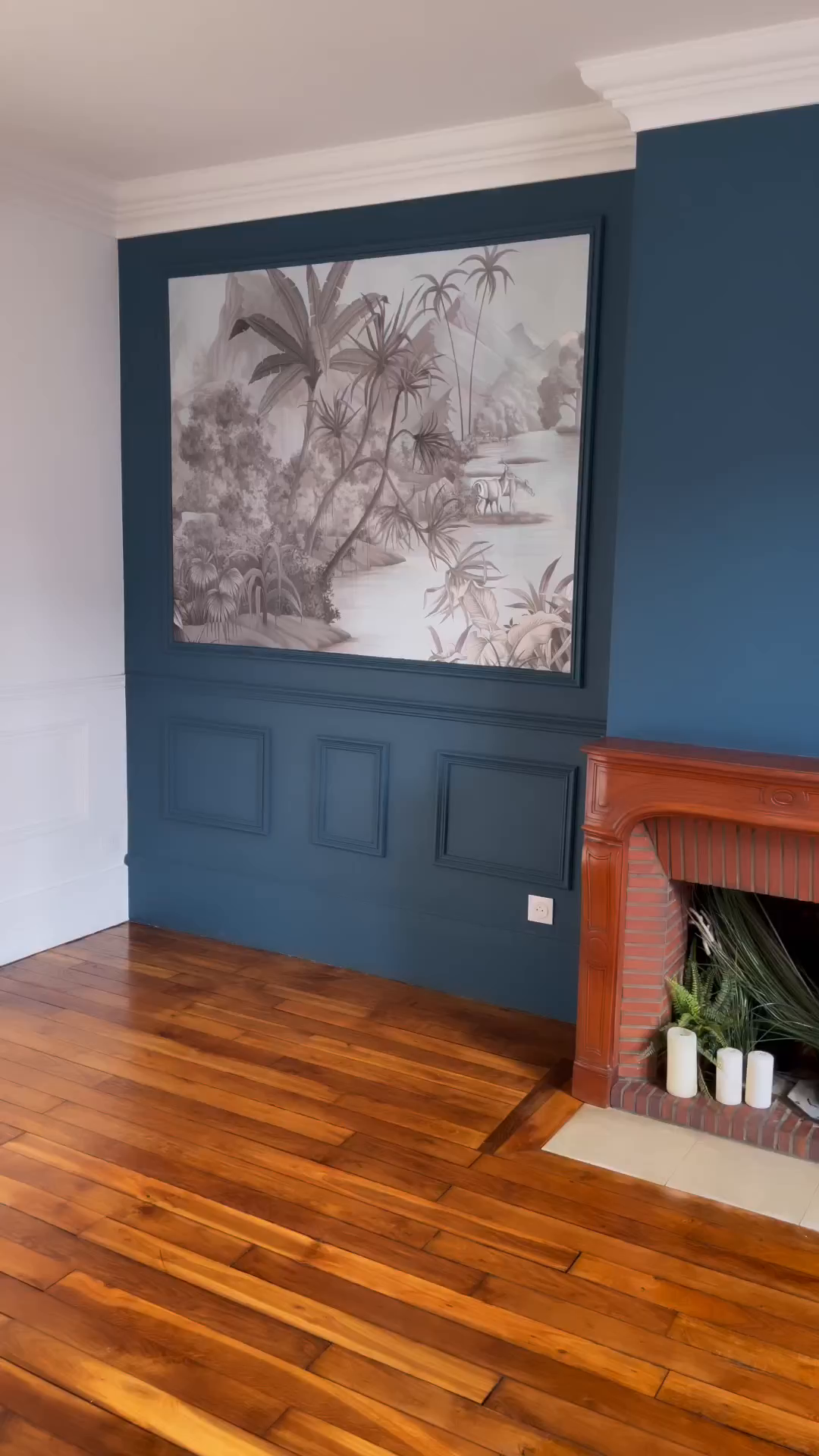 Living room with blue walls, wood floors, and a brick fireplace. A large tropical mural hangs above paneled wainscoting.