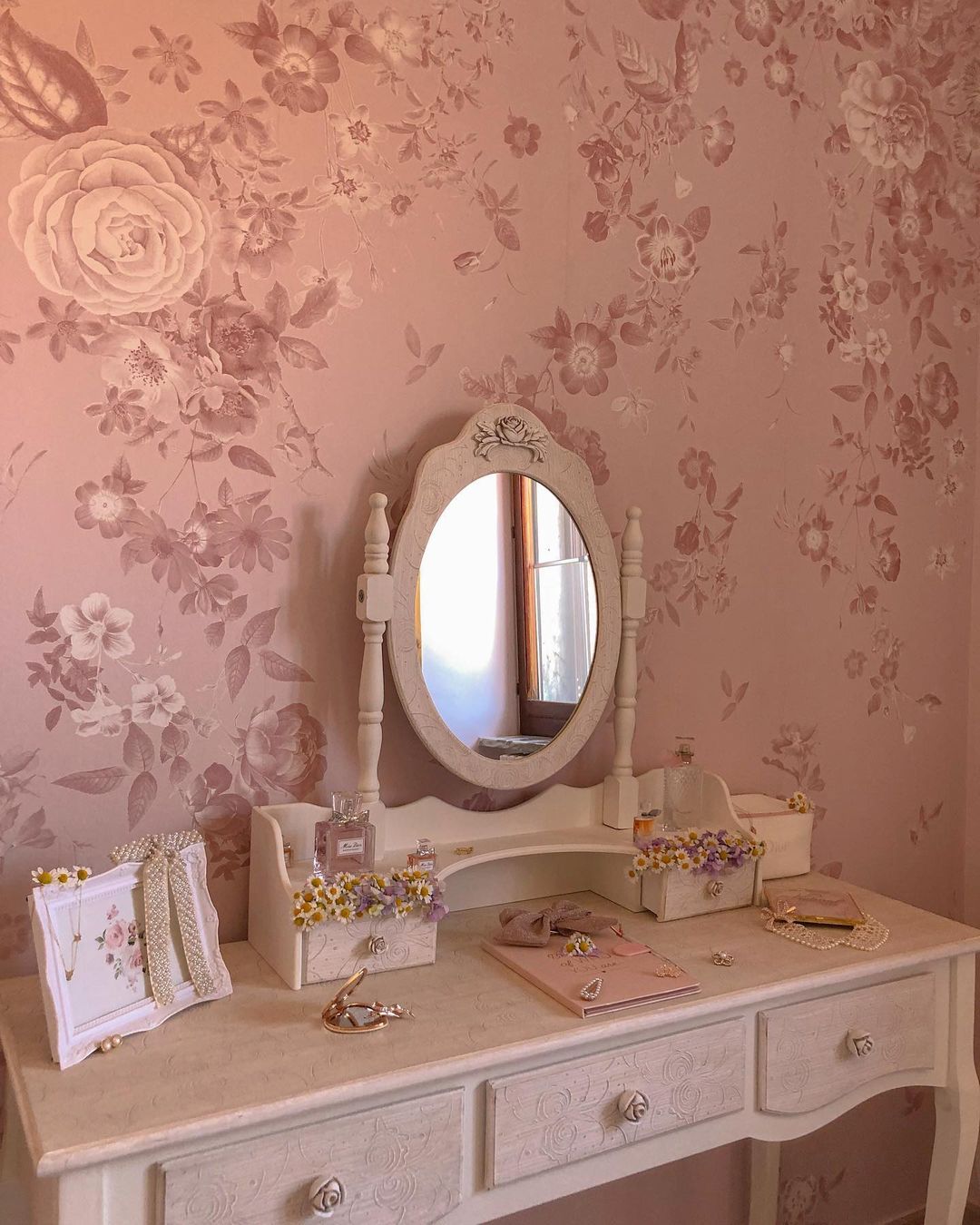 Bloemig roze behang achter een witte wastafel met ovale spiegel, parfum, sieraden en een ingelijste foto.