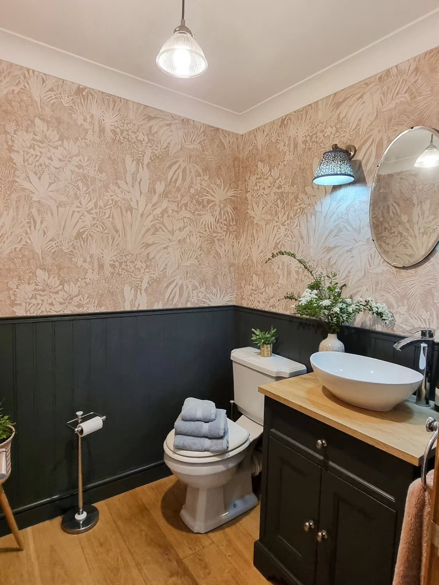 Un pequeño baño con papel tapiz botánico rosa claro encima de paneles oscuros. Se ve un inodoro blanco, un tocador de madera con un lavabo de sobre encimera blanco y un espejo ovalado sobre el suelo de madera clara.