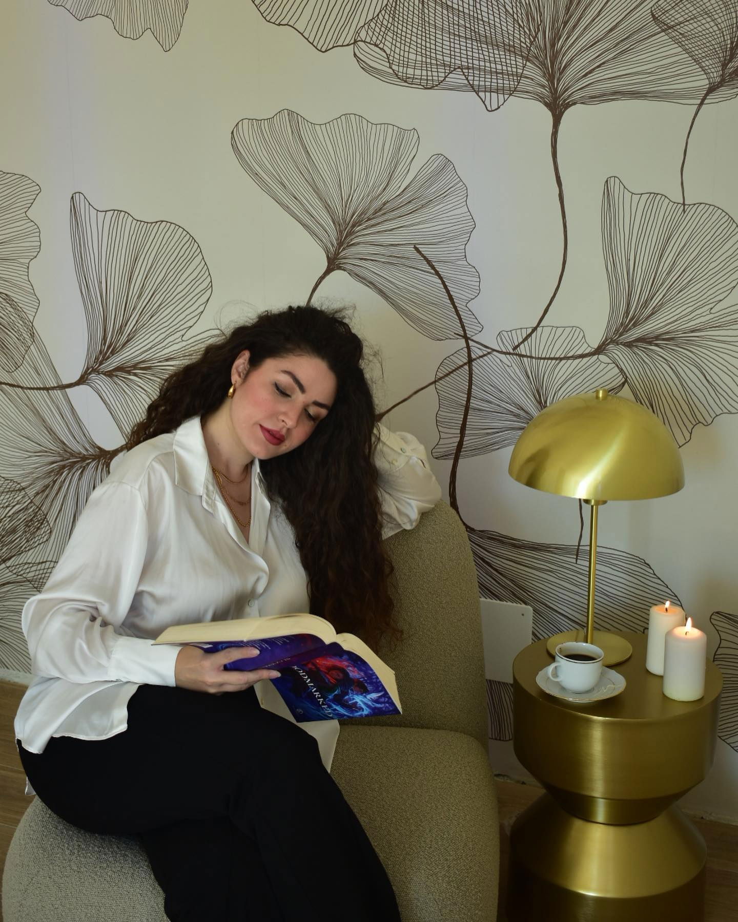 Femme lisant un livre dans un fauteuil à côté d'une table d'appoint dorée avec une lampe, un café et des bougies.