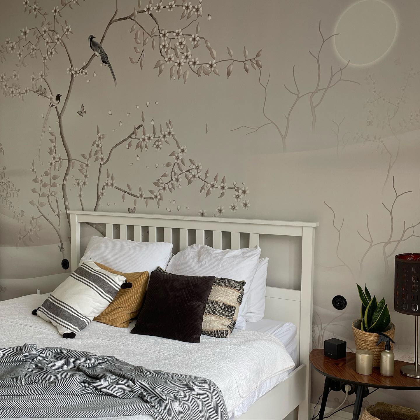 Chambre avec lit blanc, coussins à motifs et jeté à chevrons gris. L'art mural représente des oiseaux, des branches et une lune.