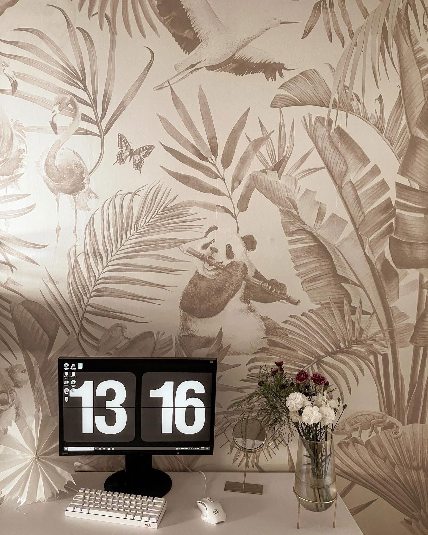 Installation de bureau avec horloge numérique, clavier, souris et fleurs sur fond de papier peint tropical.