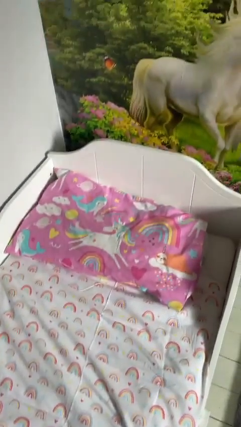 White crib with rainbow sheets and pink unicorn pillow, unicorn mural on wall.