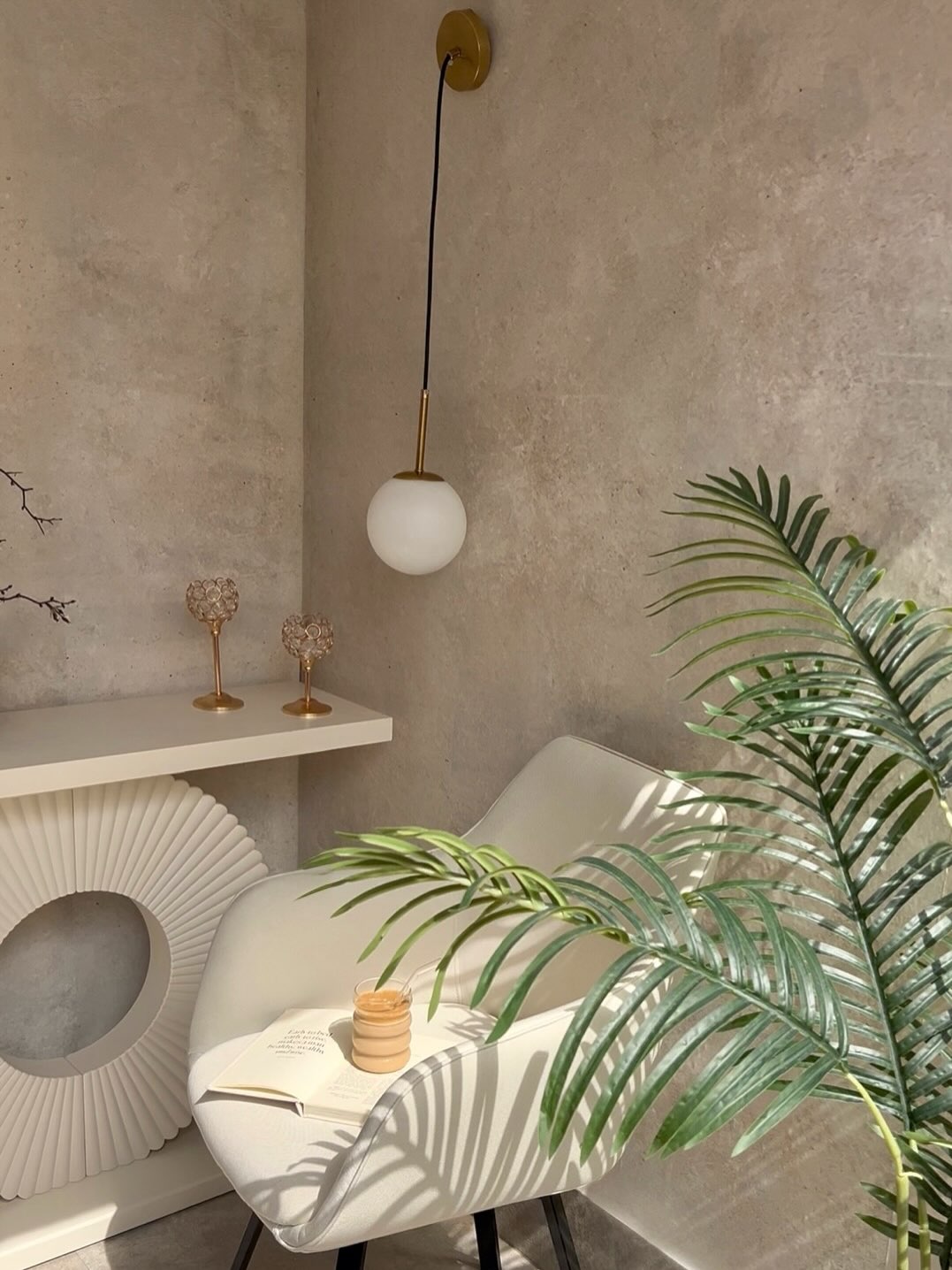 A modern gold and white globe wall light illuminates a cream chair with a book and cup, beside a large green plant, against a neutral textured wall.