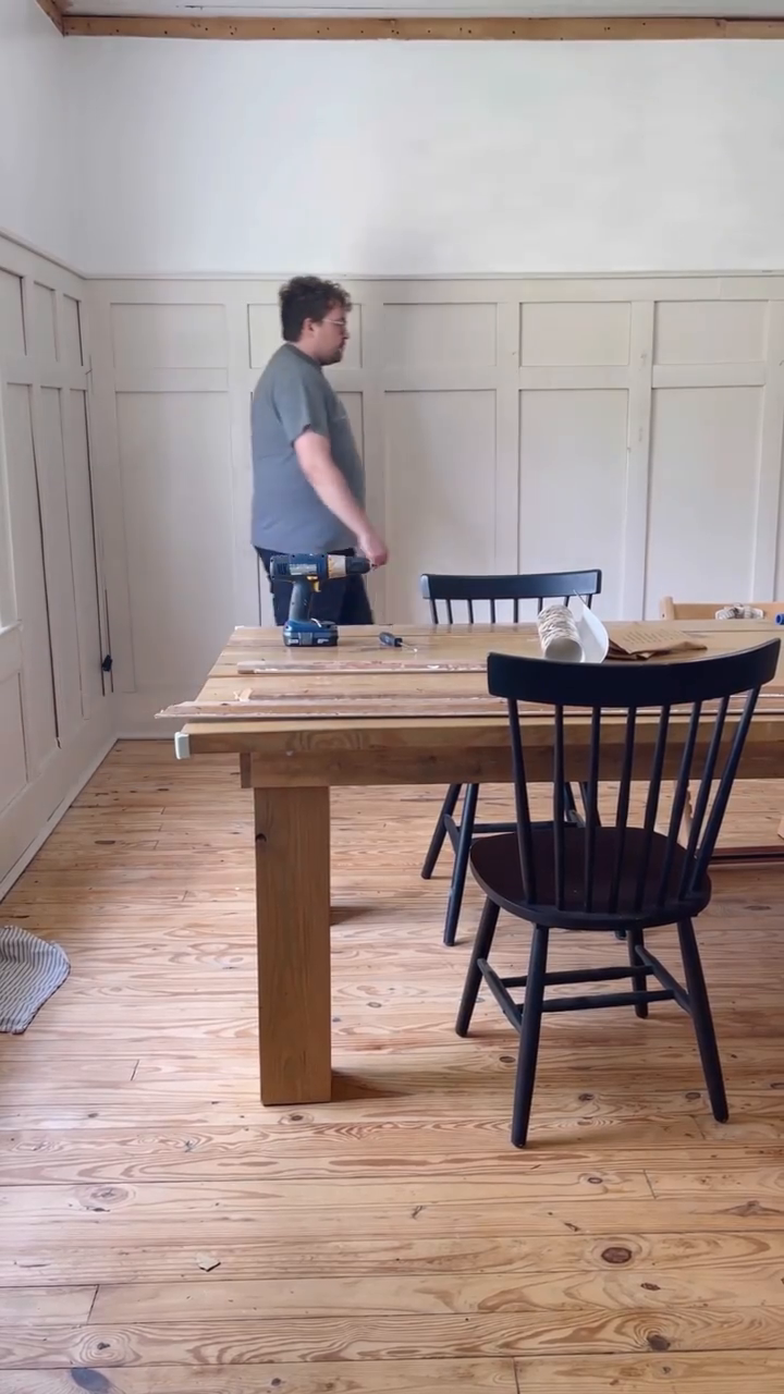 Interior shot of a man working on a wooden table with a drill in a room with paneled walls.