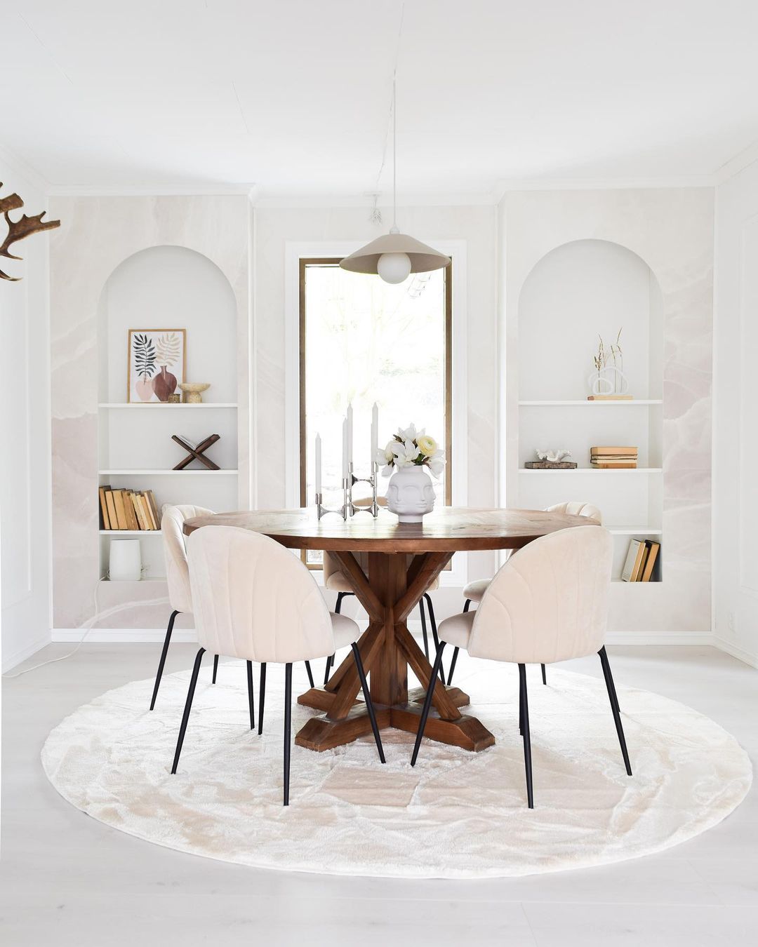 Sala da pranzo luminosa con tavolo rotondo in legno, quattro sedie color crema e scaffali a muro.