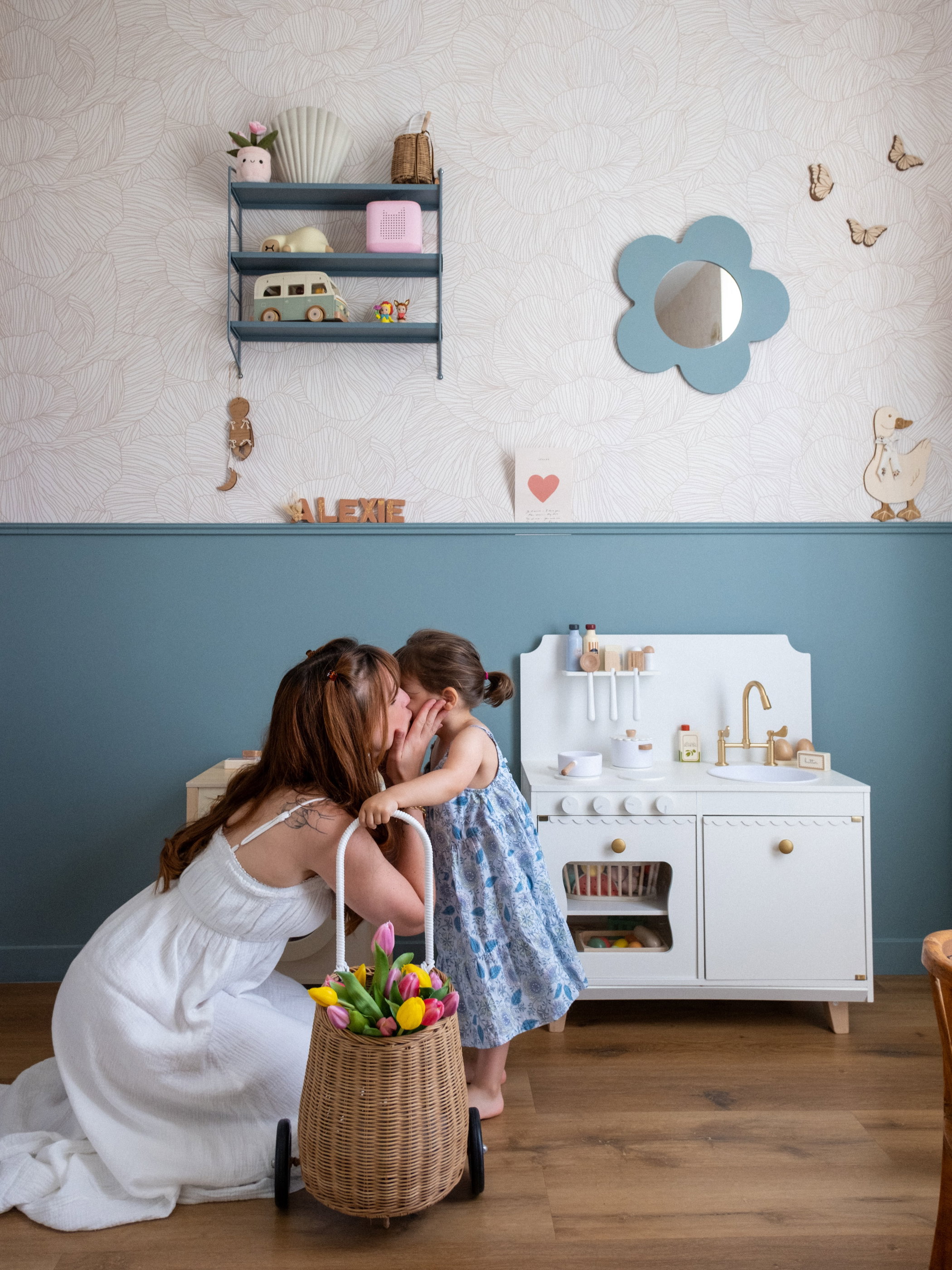 Mom kisses daughter near a white toy kitchen with a basket of tulips.