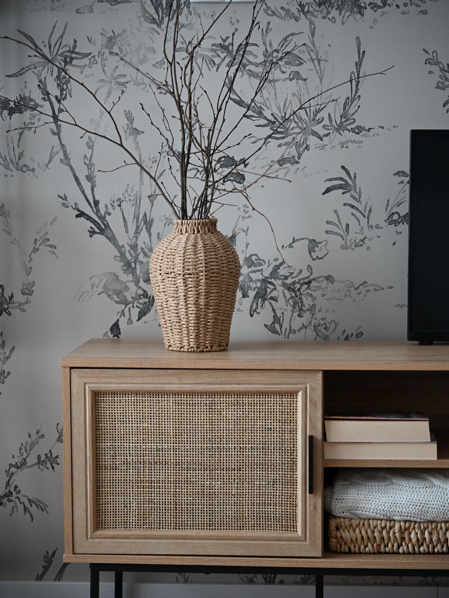 Wicker vase with branches on a wood and rattan media console against floral wallpaper.