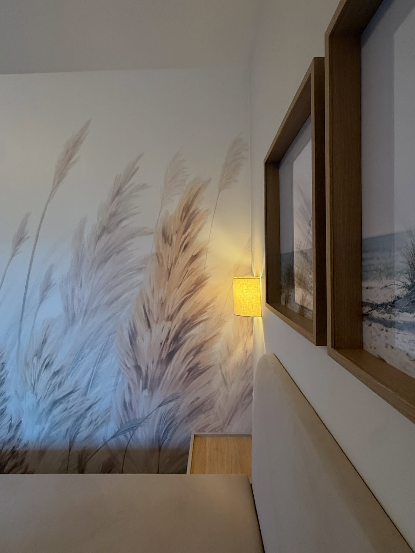Hoek van een slaapkamer met behang met tarwethema, een wandlamp en ingelijste strandkunst.