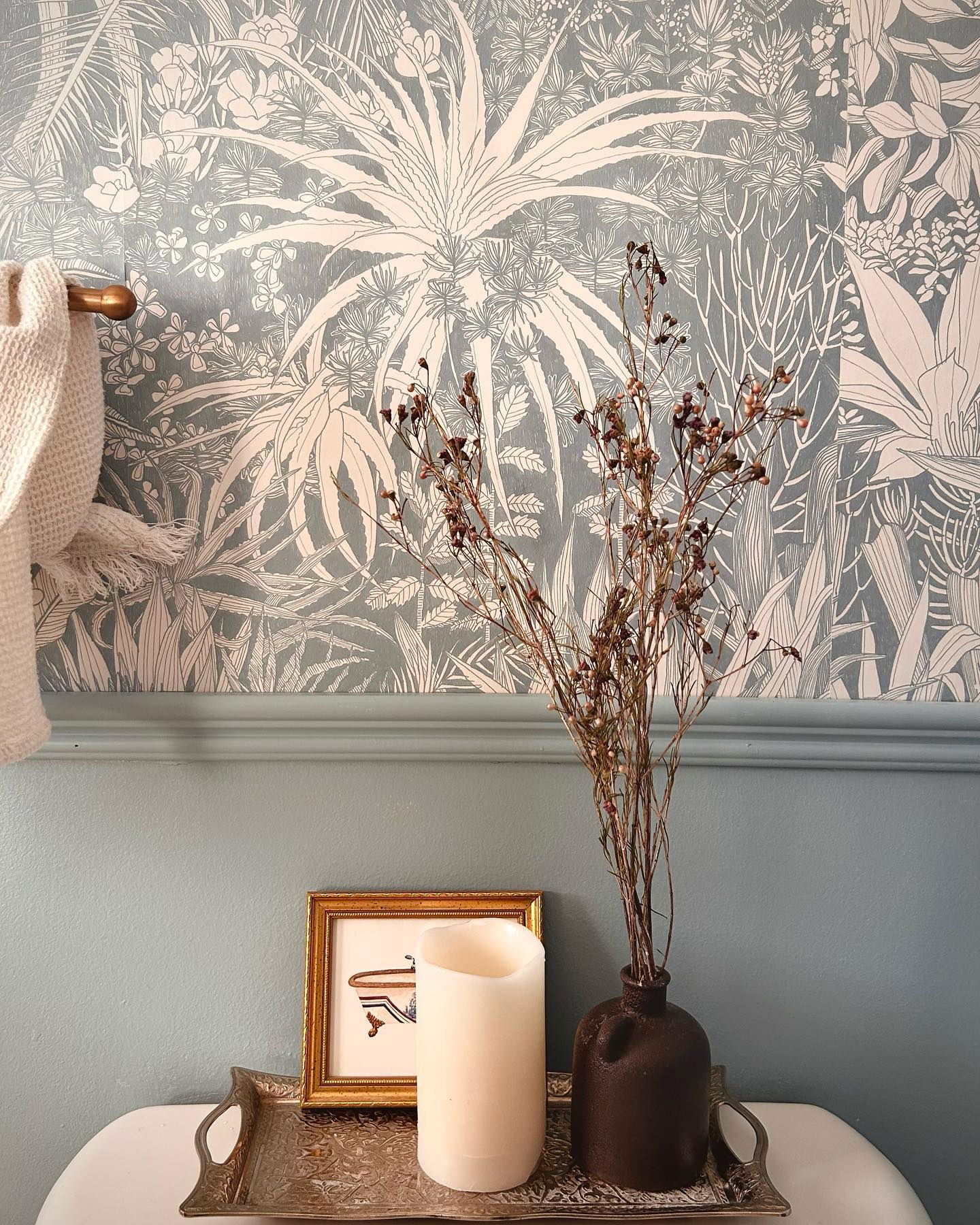 Décoration de salle de bain sur les toilettes&nbsp;: arrangement floral séché, bougie, œuvre d’art encadrée sur un plateau argenté. Papier peint botanique bleu.