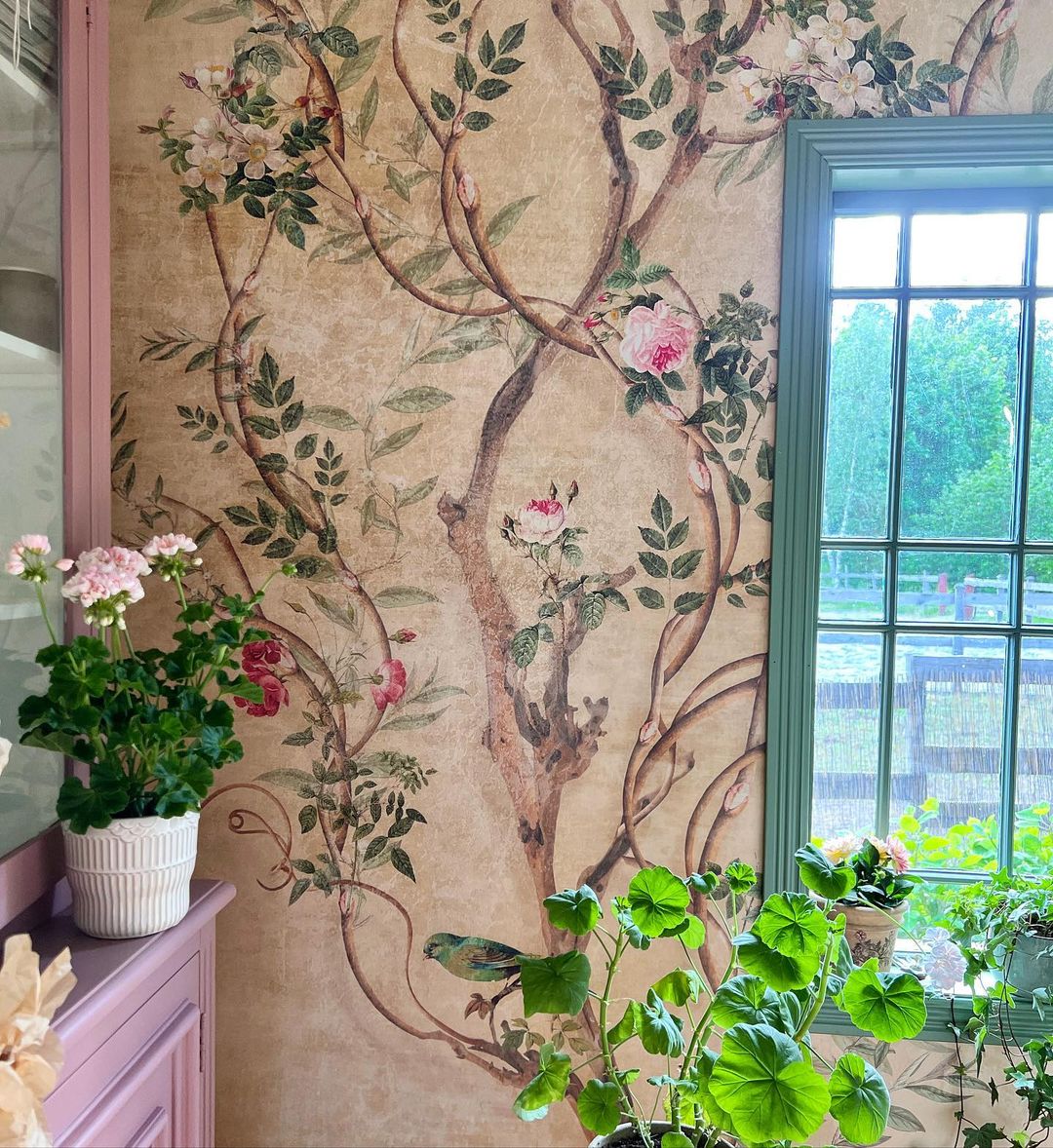 Interiør med blomstret tapet, lyserød kommode og grønt vindue. Potteplanter står på kommoden og vindueskarmen.