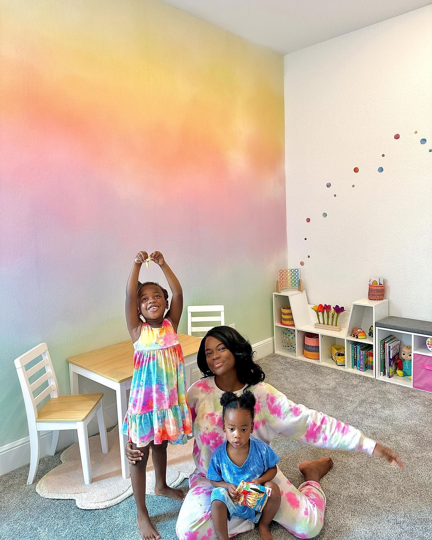 A woman and two young children pose in a playroom with colorful decor.