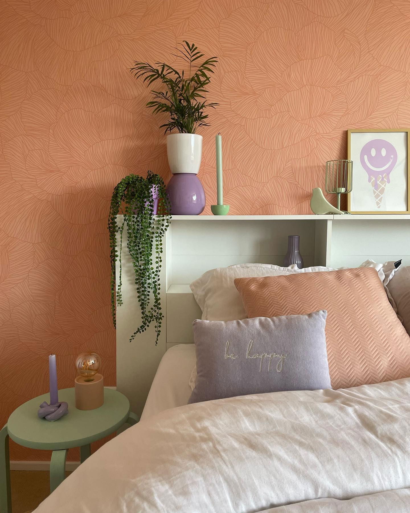 Dormitorio con papel tapiz color melocotón, estante de cabecera blanco, plantas y decoración en colores pastel. Las almohadas dicen "sé feliz".