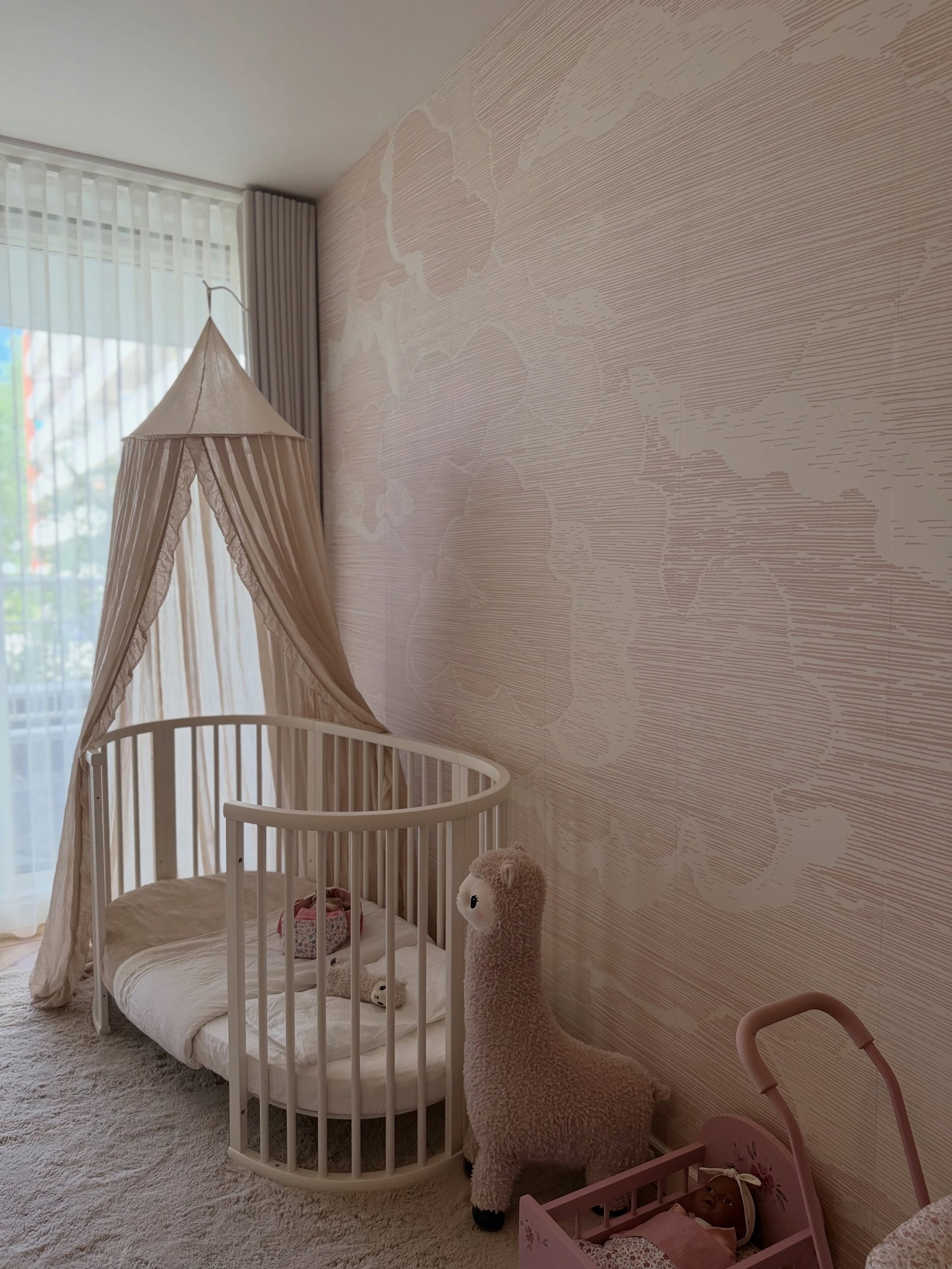 A nursery with a white oval crib, a tan canopy, and a fluffy pink llama toy.