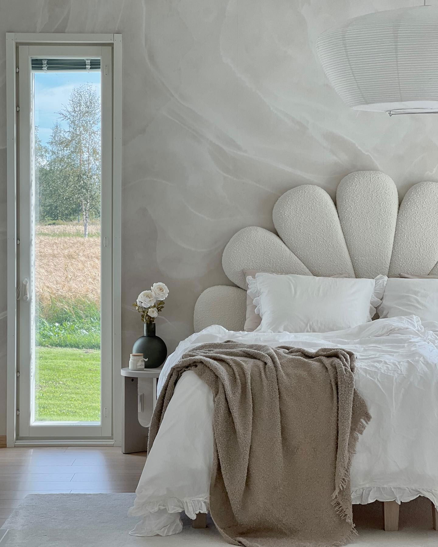 Camera da letto con testiera bianca a fiori, lenzuola bianche e coperta beige. Vista dalla finestra sul campo verde.