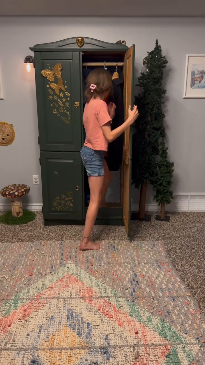 A girl opens a green wardrobe with gold butterfly decals in a whimsical room.
