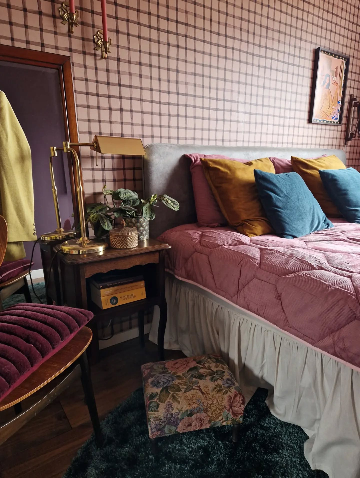 Roze gewatteerd bed met grijze hoofdsteun, gele en blauwe kussens. Roze geruite behang, nachtkastje met messing lamp, wijnrode stoel en voetenbank op donker tapijt.