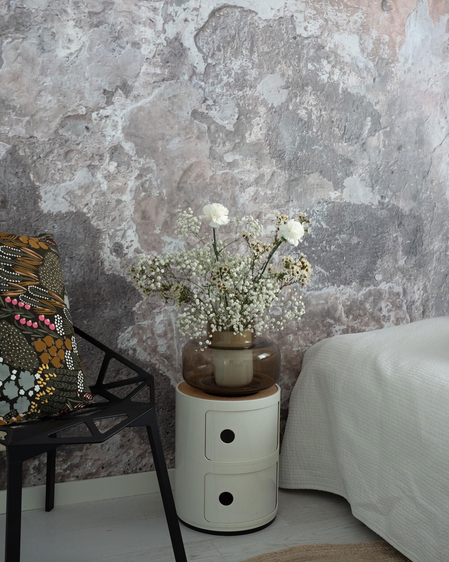 Bedroom corner with flowers, chair, and Kartell Componibili nightstand against a textured wall.