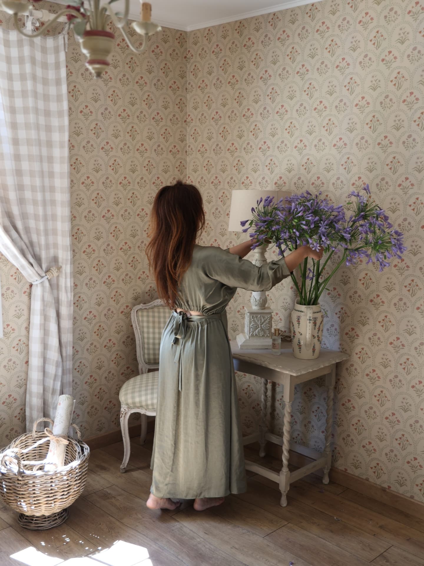 Woman arranging purple flowers in a vase on a table in a patterned room.