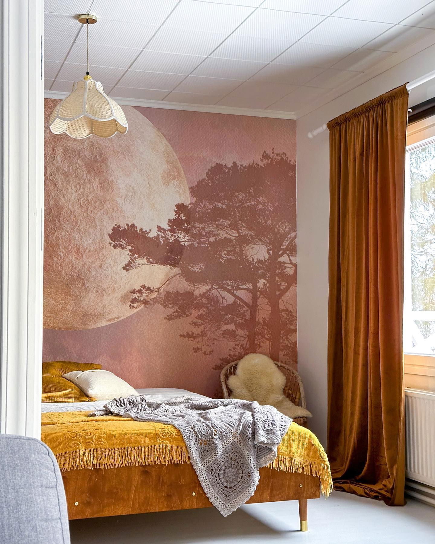 Bedroom with moon and tree wallpaper, yellow bedspread, and rattan pendant light.