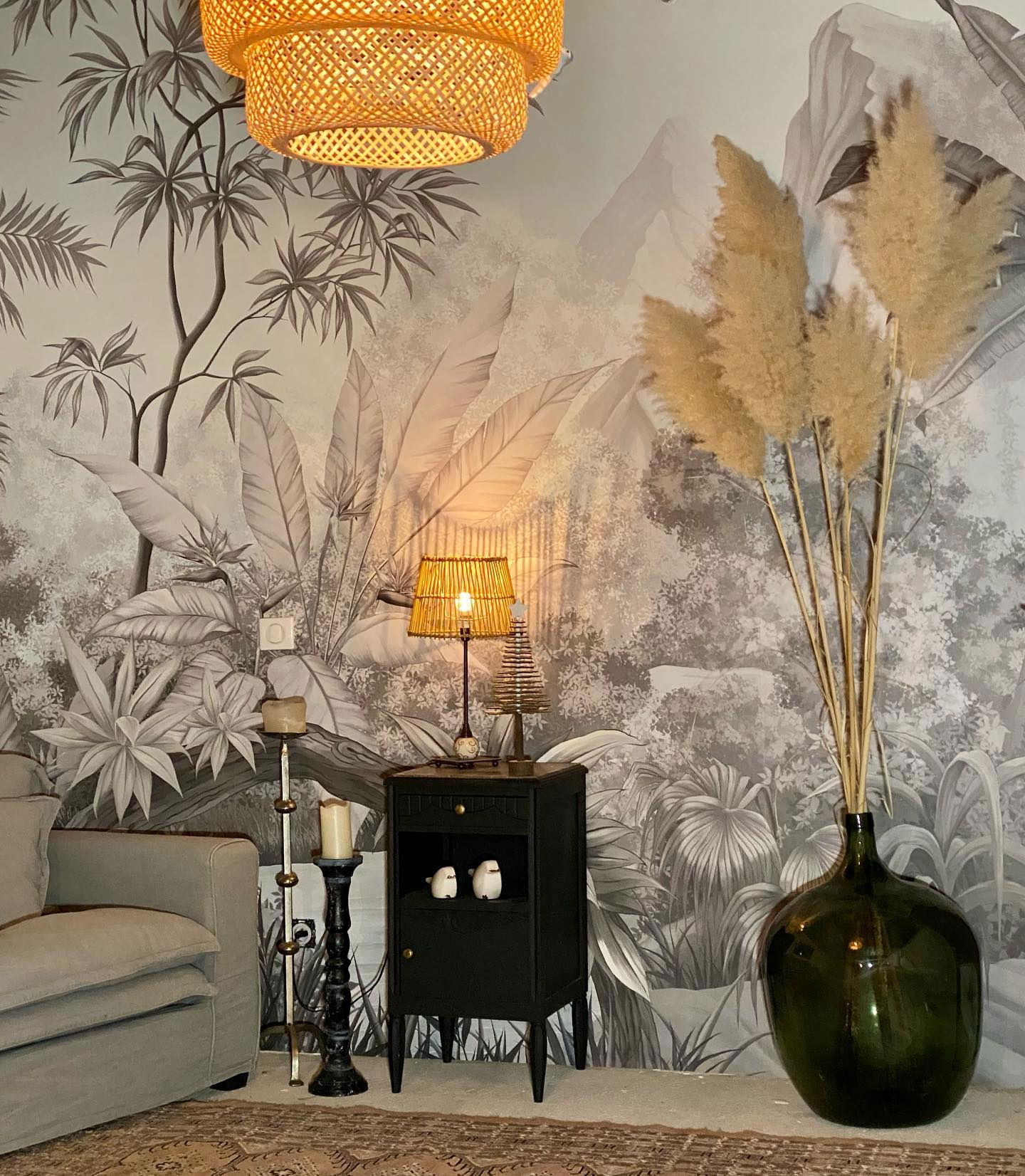 Living room scene with tropical wallpaper, rattan light fixture, and pampas grass in a green glass vase.