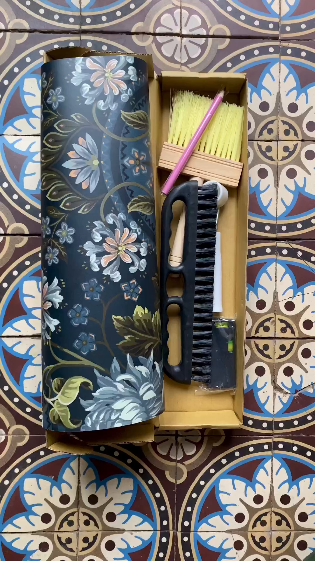 Tile stenciling kit with floral-patterned container, brushes, and level on patterned tile.