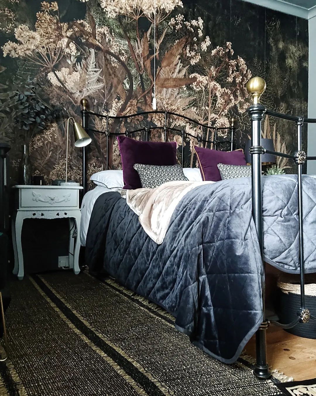 Schlafzimmer mit schwarzem Metallbett, Blumentapete und grauer Samtdecke. Nachttisch mit Lampe und gestreiftem Teppich sichtbar.
