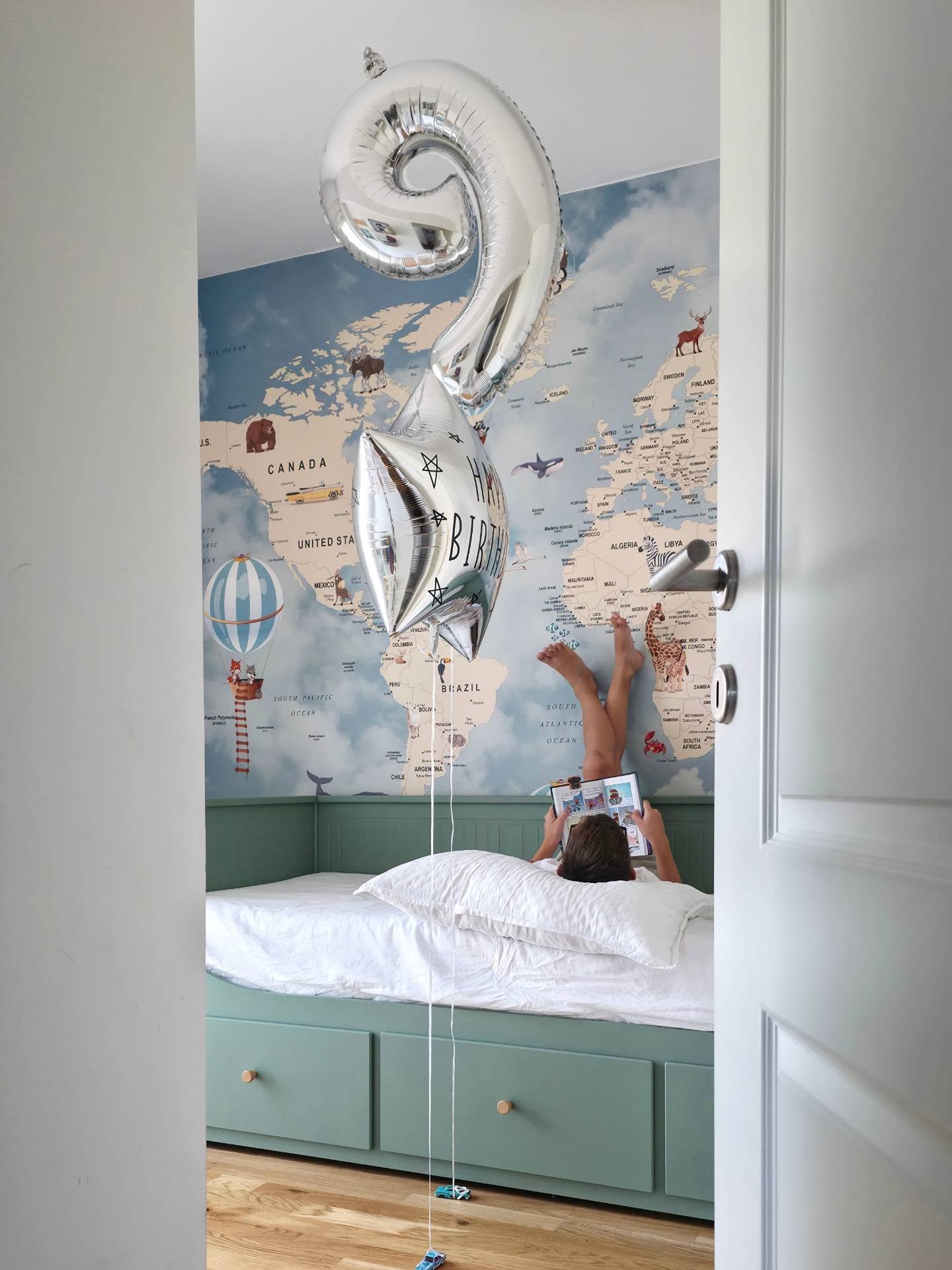 A child lies upside down on a teal bed, looking at a tablet. Silver '9' and 'Happy Birthday' balloons float above. A world map mural is on the wall.