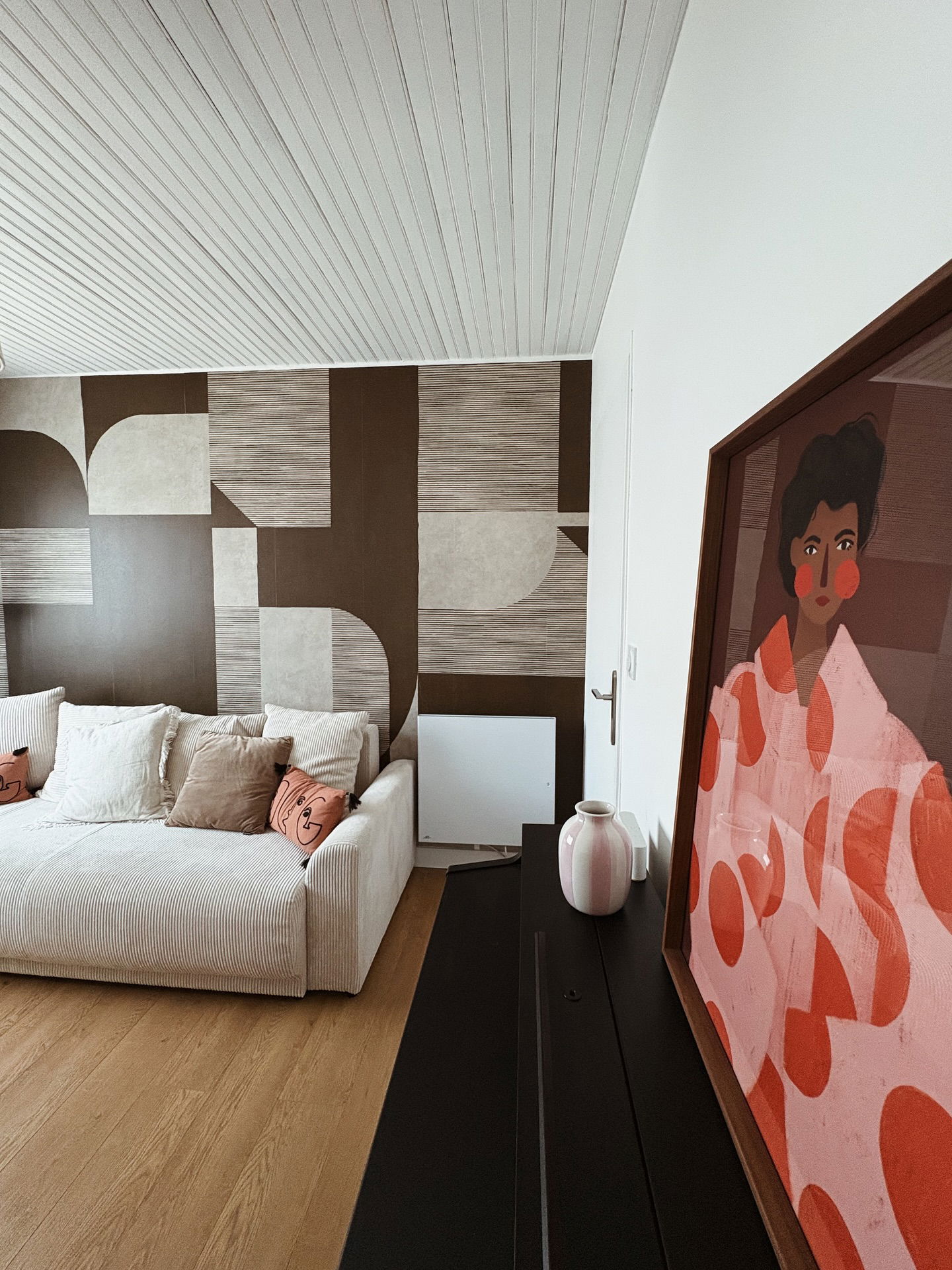 Sofa with patterned wallpaper and framed art on a dark cabinet with a pink vase.