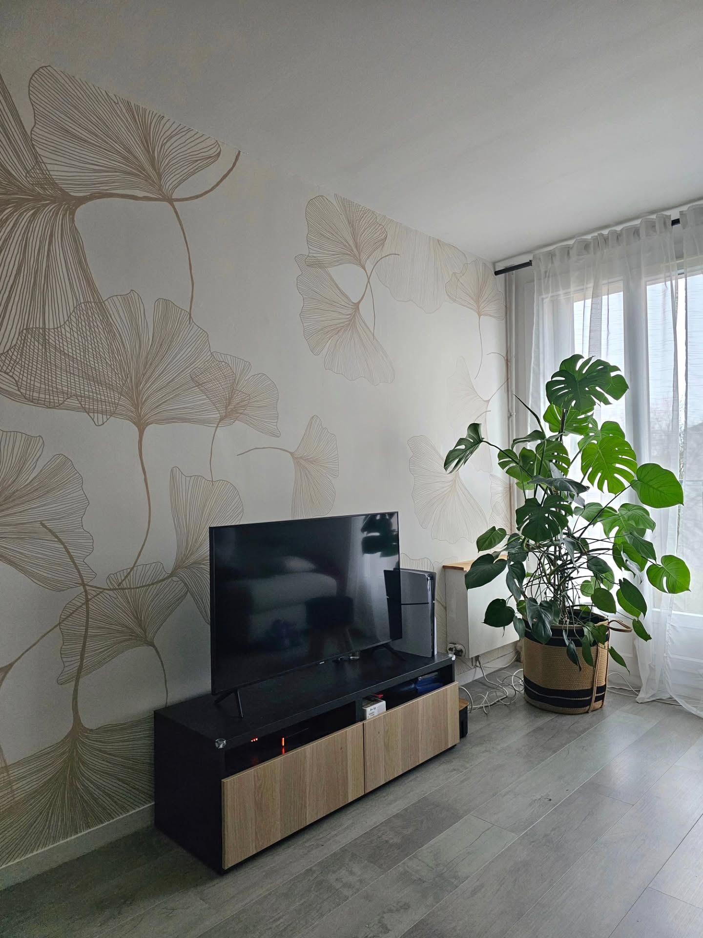 Nowoczesny salon z dużą rośliną, telewizorem na drewnianej szafce i tapetą z liśćmi ginkgo.