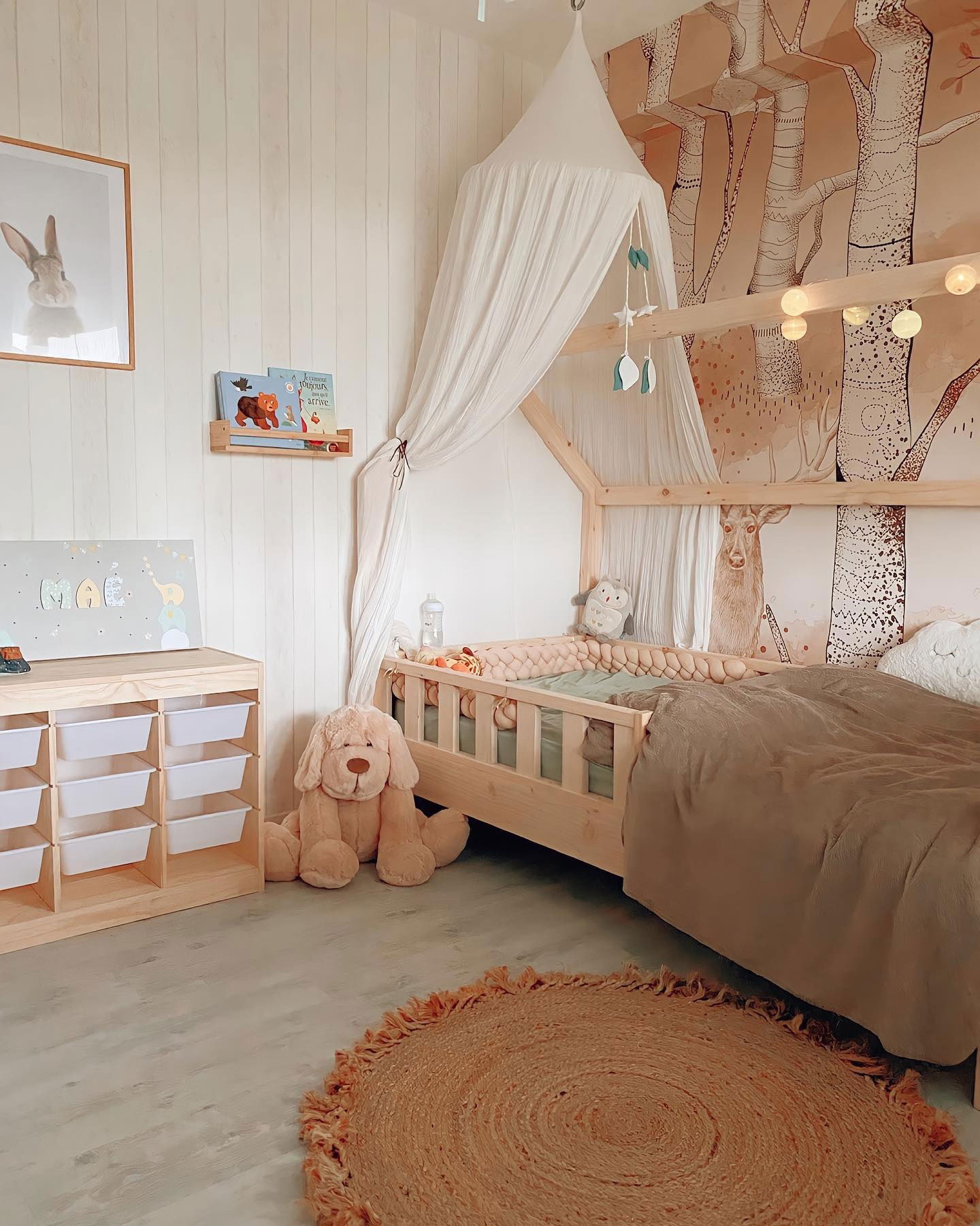 Kinderzimmer mit hölzernem Bettgestell, Baldachin und Waldgemälde. Ein Stoffhund sitzt in der Nähe einer Aufbewahrungseinheit.