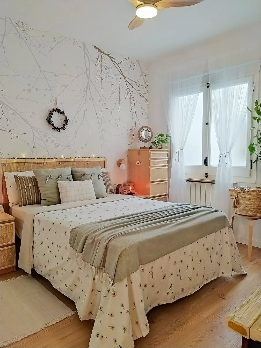 Bedroom with a rattan headboard bed, light floral bedding, and a green throw. A wall mural of tree branches and a dark wreath are behind it.