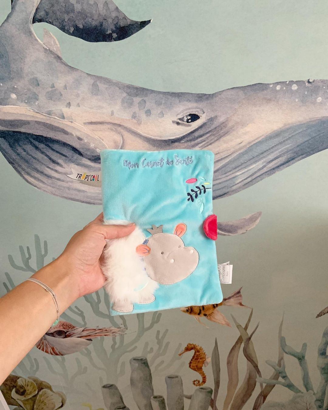 Hand holding a blue plush baby book with a hippo appliqué, against an ocean-themed wallpaper.