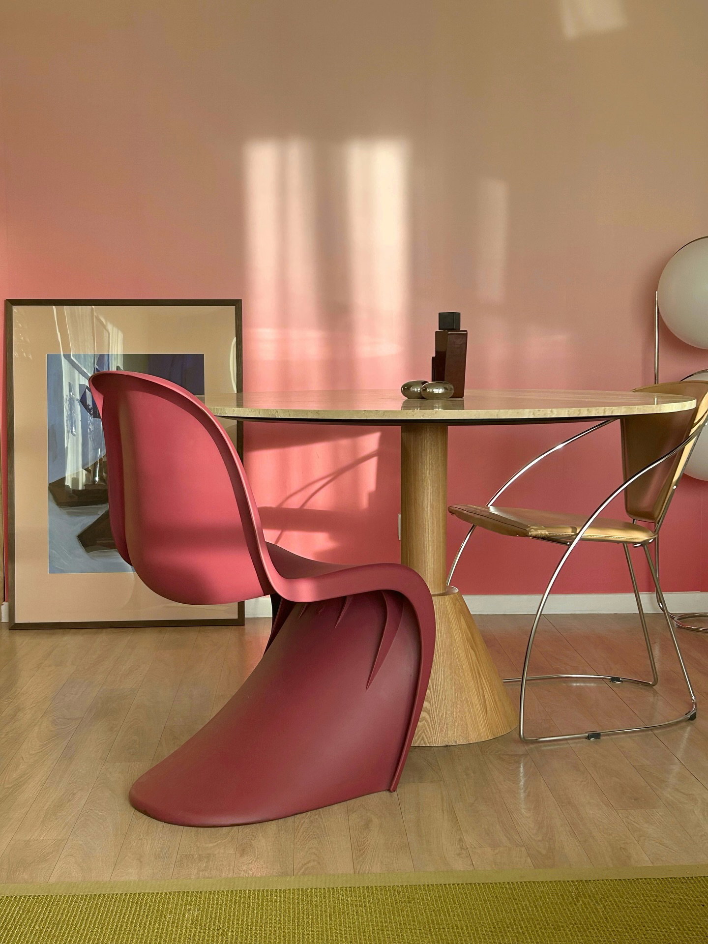 Ein geschwungener, dunkelpinker Stuhl und ein runder, hellholzfarbener Tisch in einem modernen Raum. Die Wand hat einen pinken Farbverlauf mit Sonnenlichtschatten, und ein grüner Teppich liegt auf dem Boden.