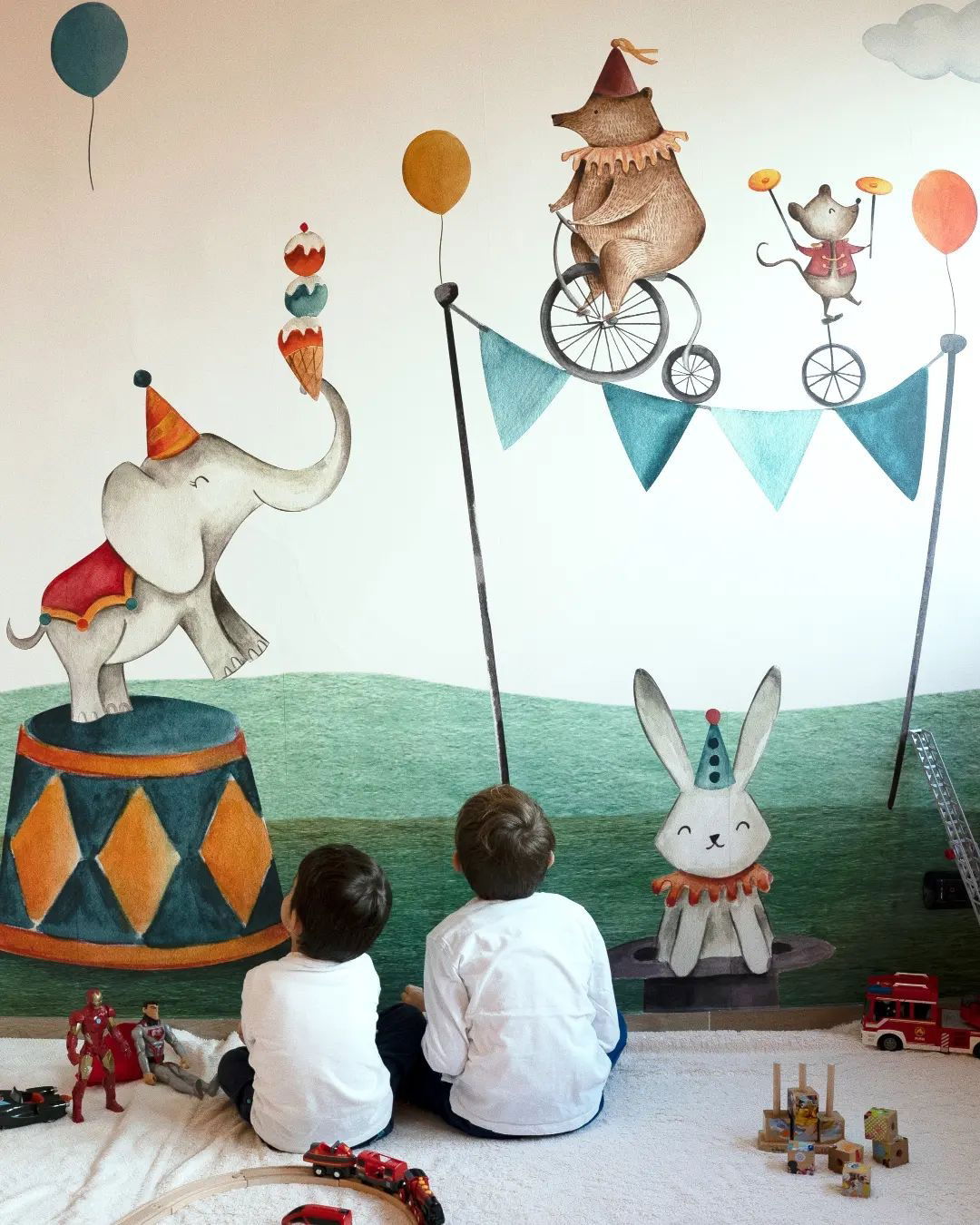 Two boys sit before a circus-themed mural with an elephant, bear, and rabbit, surrounded by toys.