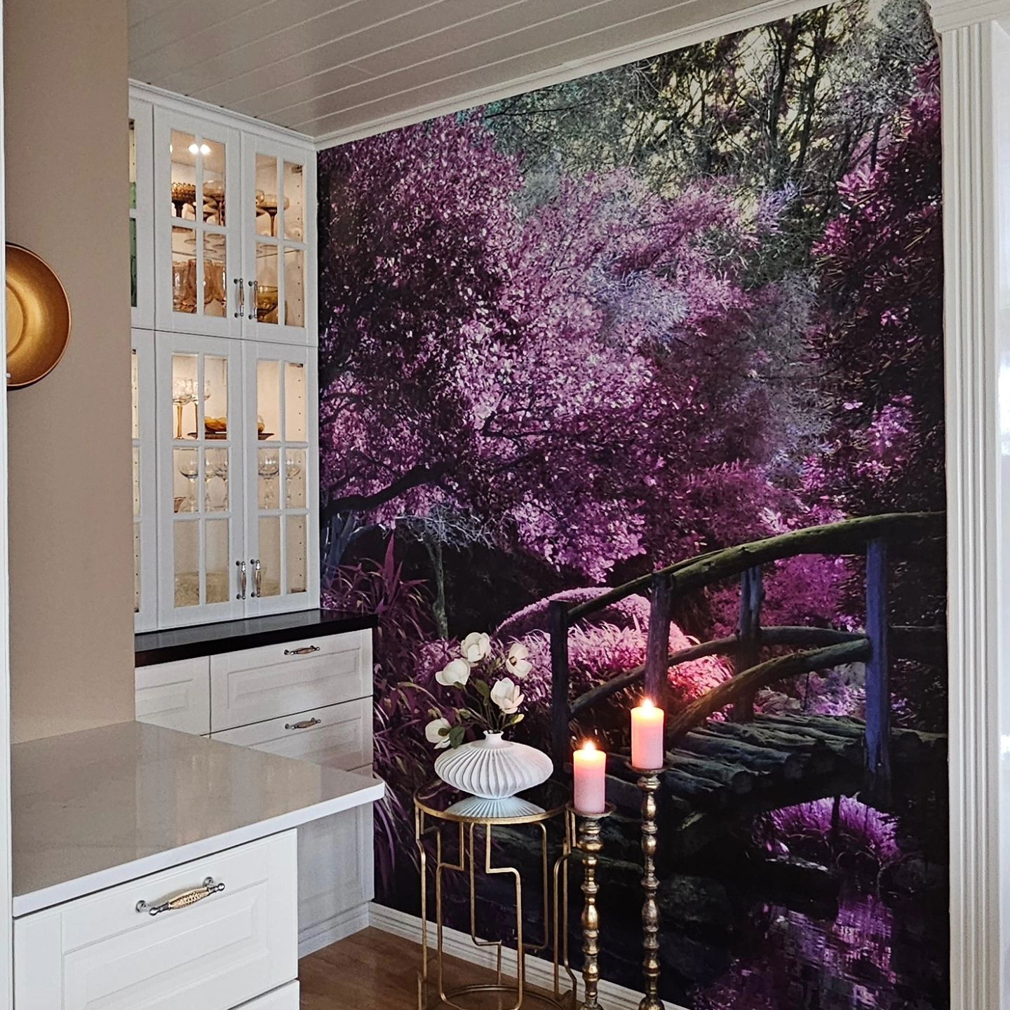 Interieur mit lila Waldwandbild, Brücke und Kerzen. Weiße Schränke und goldene Akzente sorgen für Eleganz.
