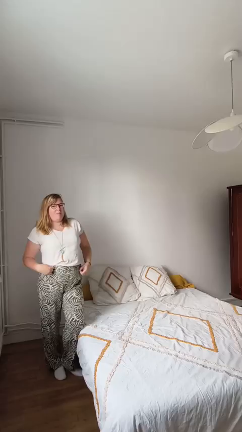 A woman in a bedroom wearing patterned pants and a white t-shirt.