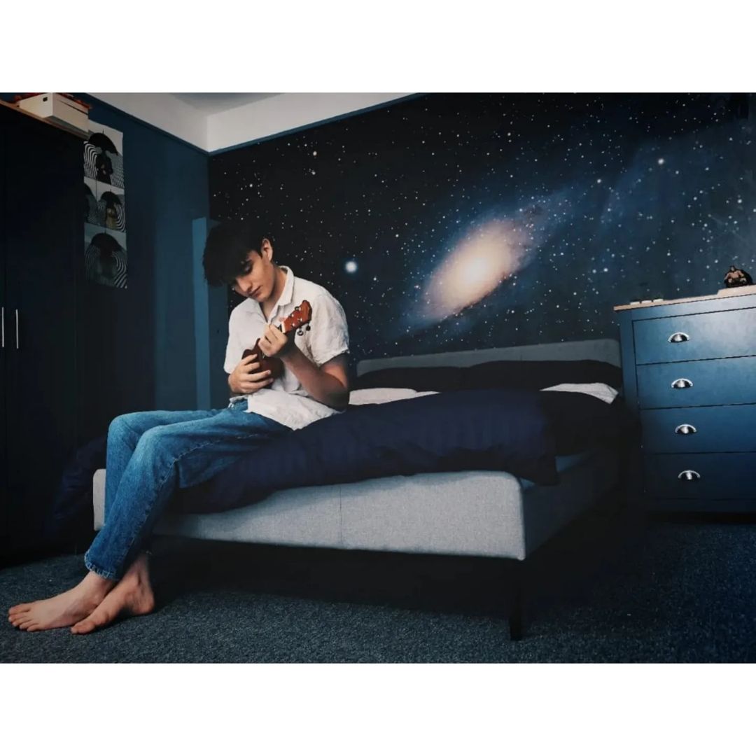 Un joven se sienta descalzo en una cama, tocando un ukelele. Fondo de pantalla de galaxia.