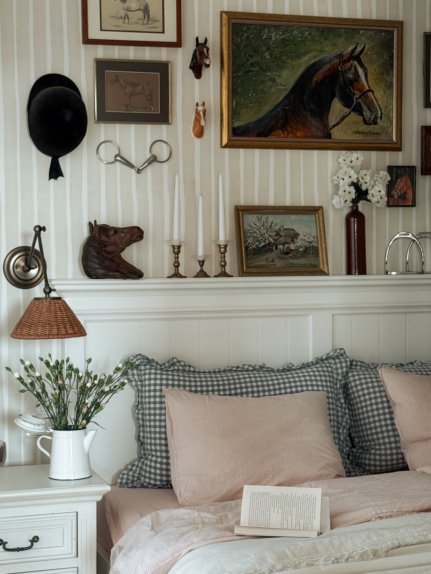 Säng med rosa och grårutiga kuddar. Ovanför en vit sänggavel, en hylla med ryttarinredning: hästmålningar, hjälm och träns.