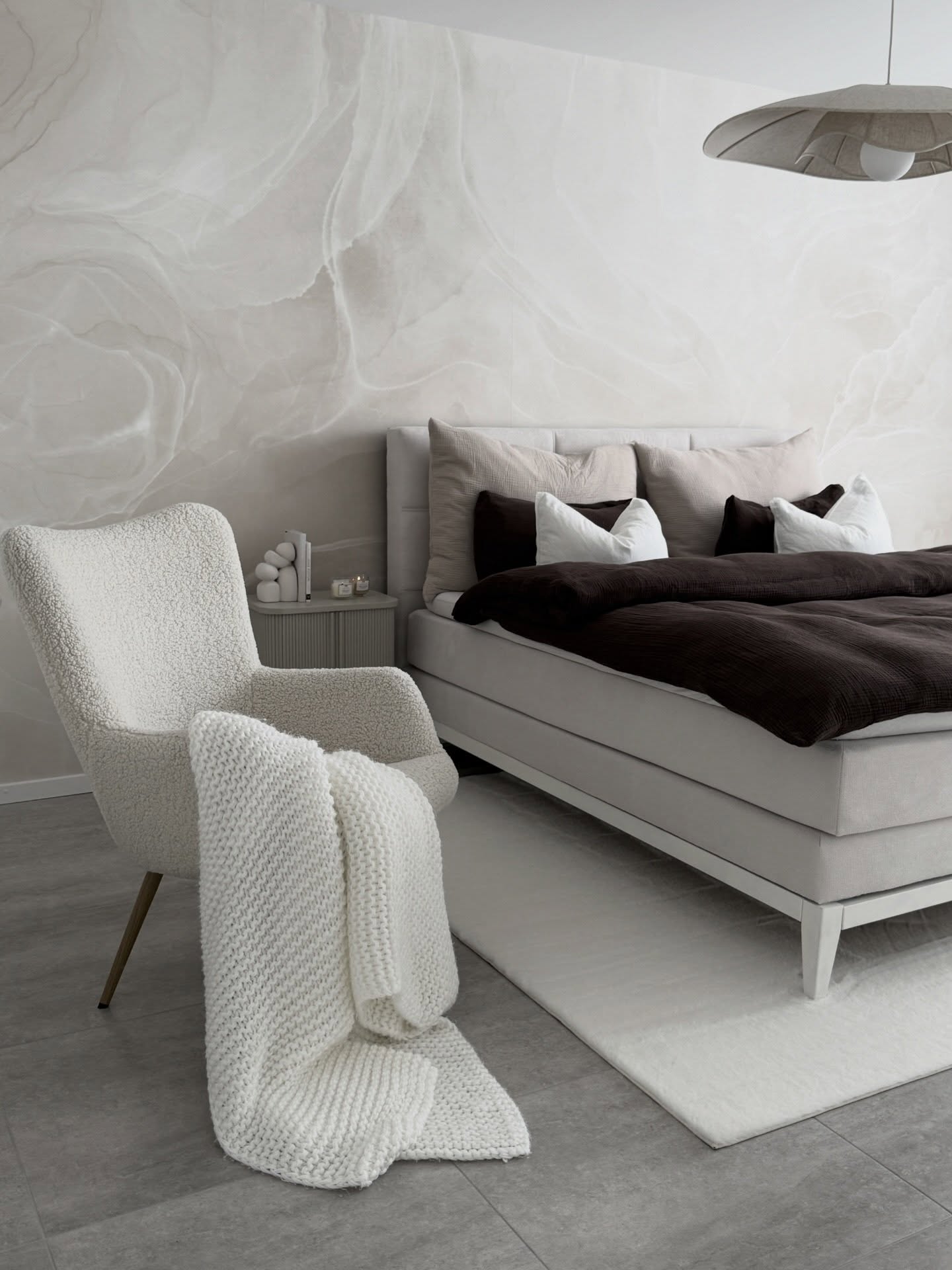 Chambre avec un lit rembourré gris, une couette marron foncé et un fauteuil blanc moelleux tenant un jeté en tricot. Le mur présente un motif abstrait clair.
