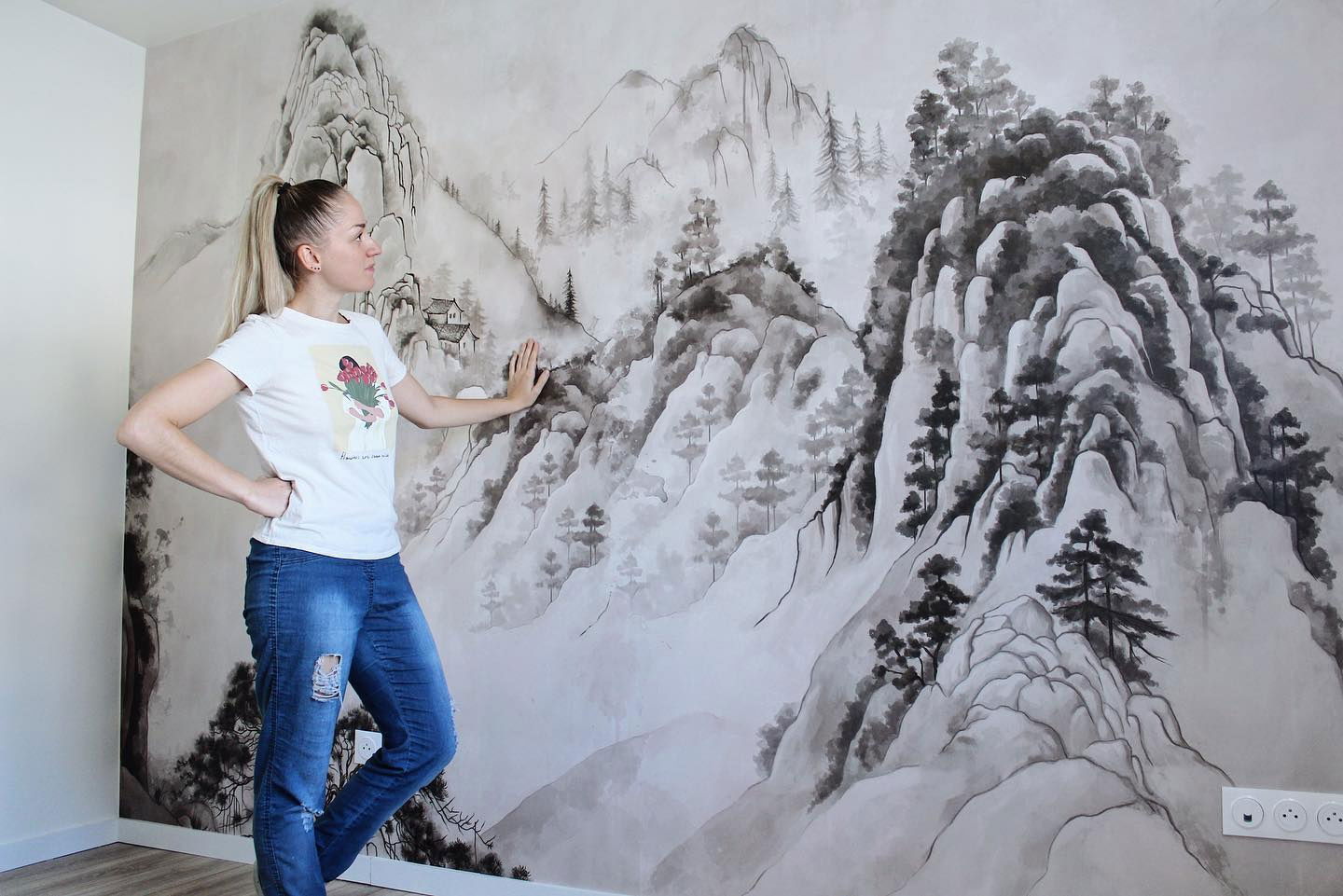 Woman in jeans and t-shirt stands in front of a wall mural of a mountain landscape.