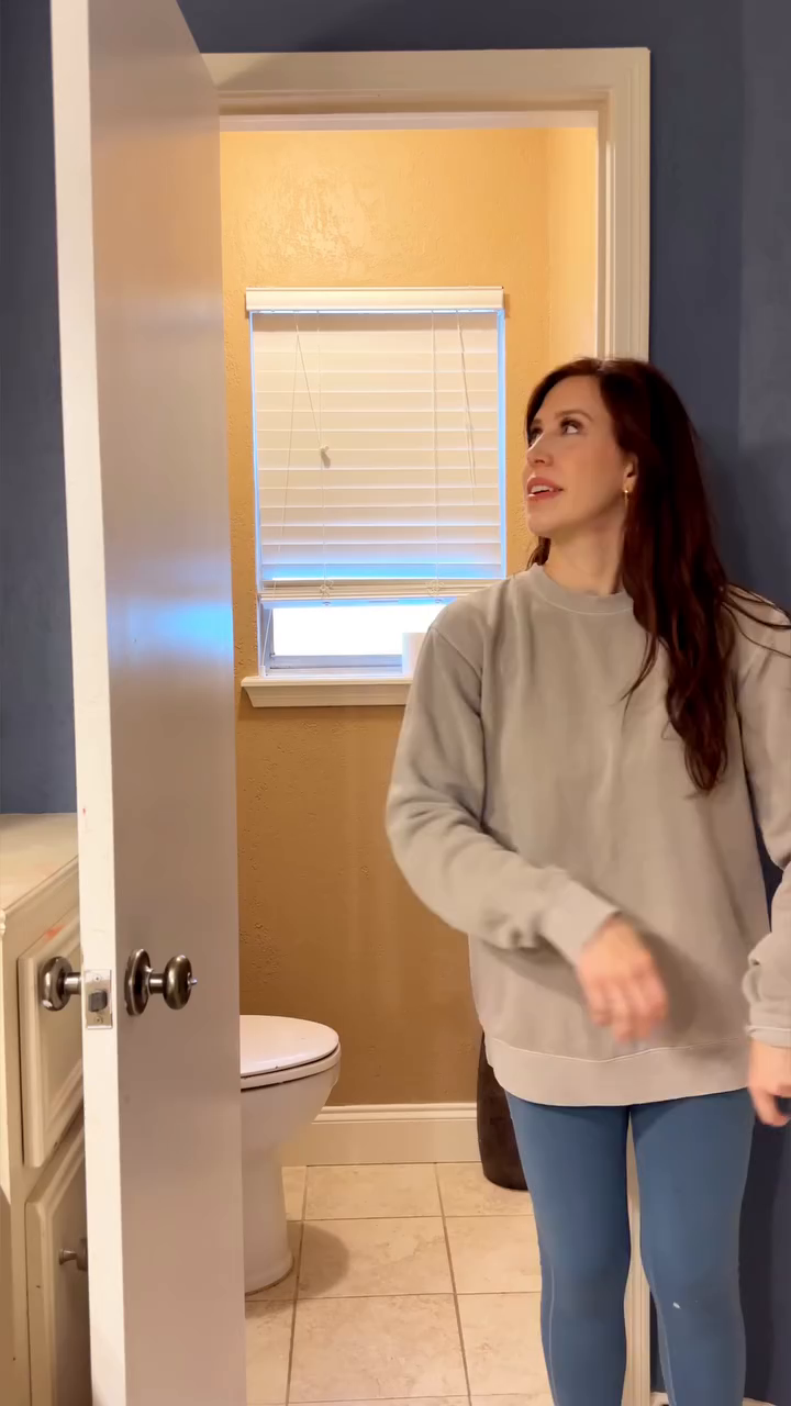 A woman in a gray sweatshirt and blue leggings stands in a blue bathroom, looking into an adjacent yellow bathroom.