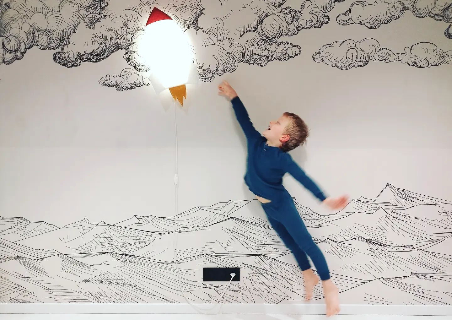 A child in blue pajamas leaps toward a rocket-shaped wall light on a wall with cloud and mountain drawings.