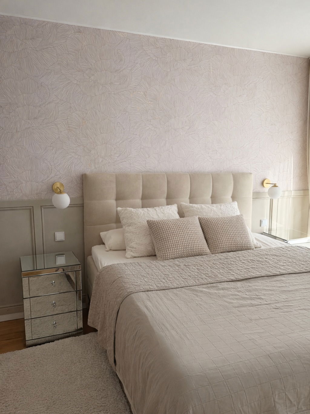A beige upholstered headboard with tufted details above a bed with quilted beige bedding and decorative pillows.