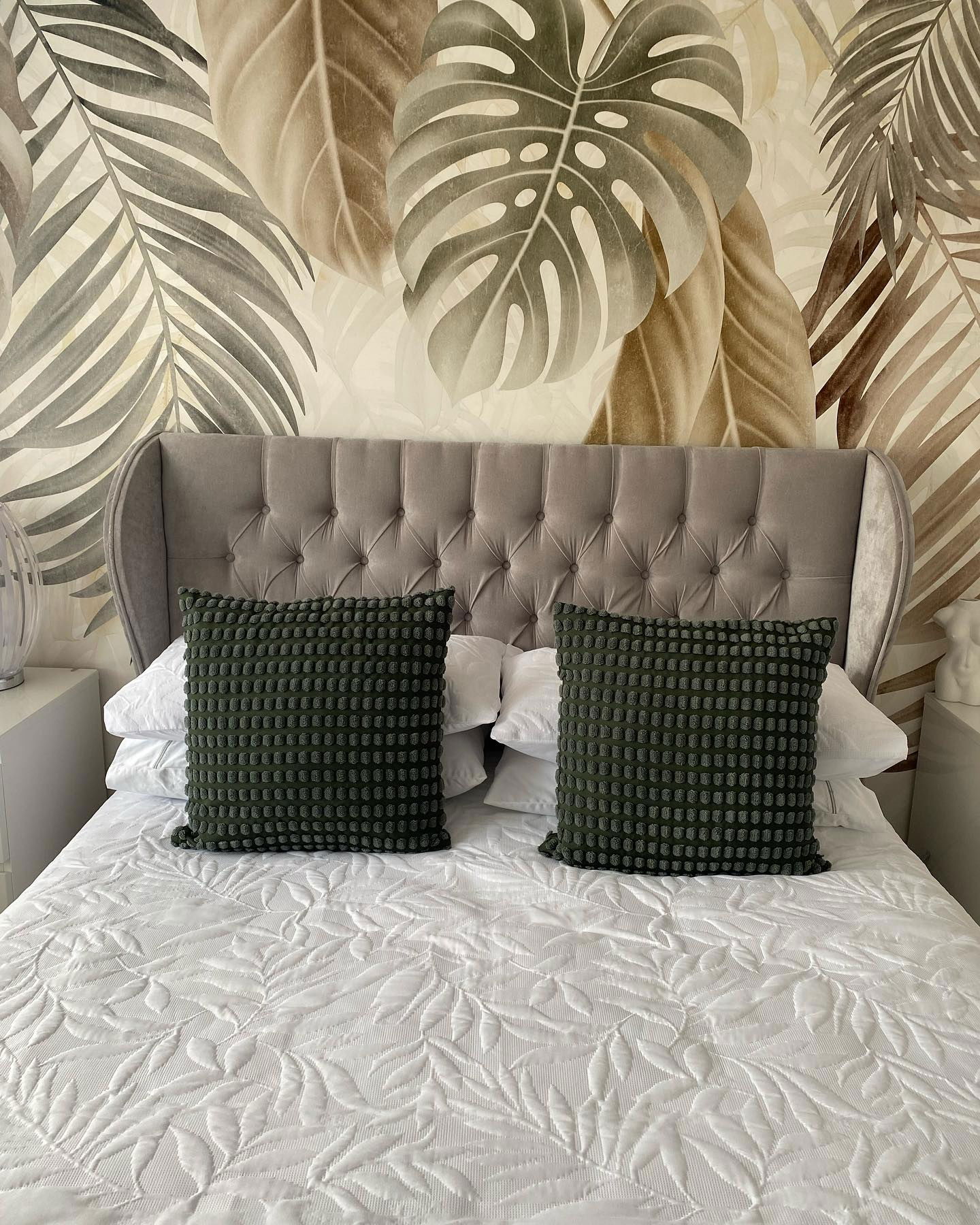 Schlafzimmer mit grauem Kopfteil, weißer Steppdecke mit Blattmuster und zwei strukturierten dunkelgrünen Kissen.