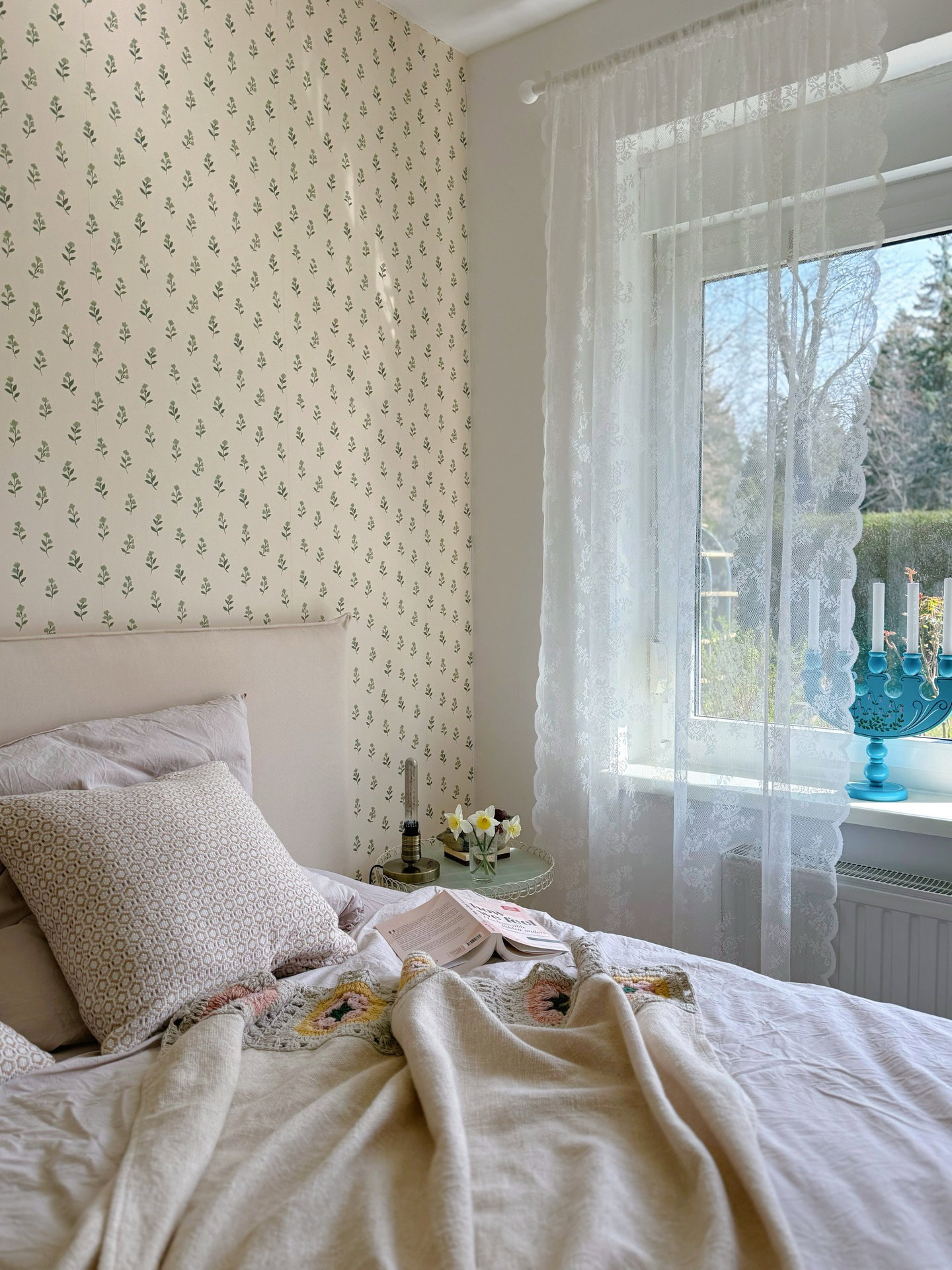 Bedroom with floral wallpaper, white lace curtains, and a bed with a crocheted throw.