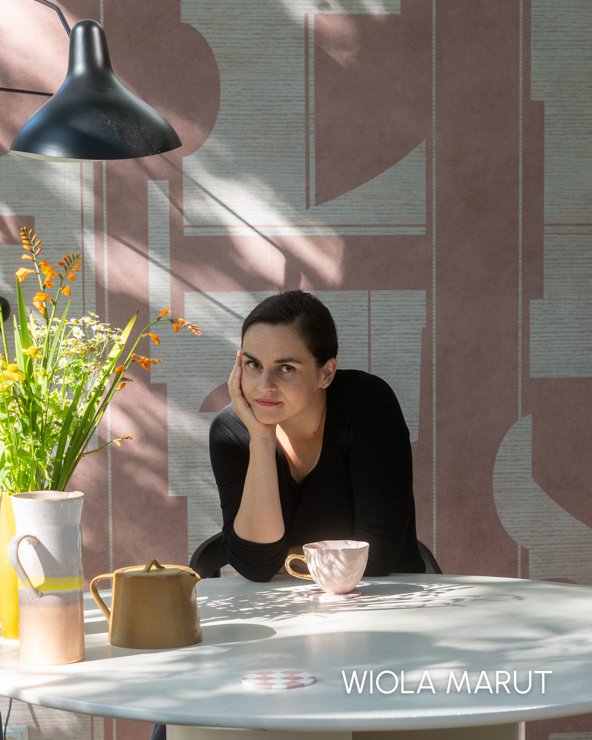 Portret van Wiola Marut aan een tafel met bloemen, thee en geometrisch behang.