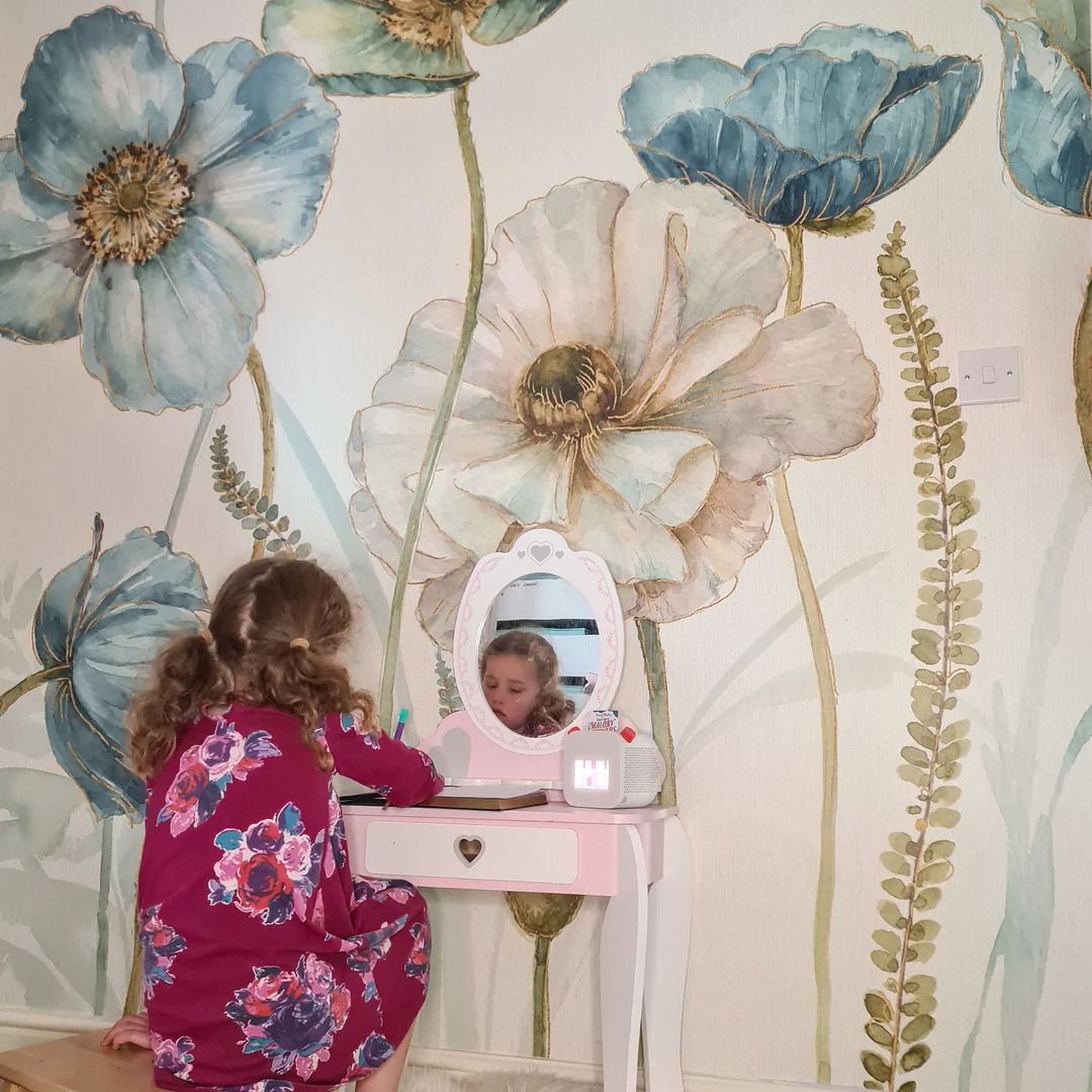 En ung jente sitter ved et rosa sminkebord med en hjertedetalj og skriver i en notatbok foran et blomstertapet.