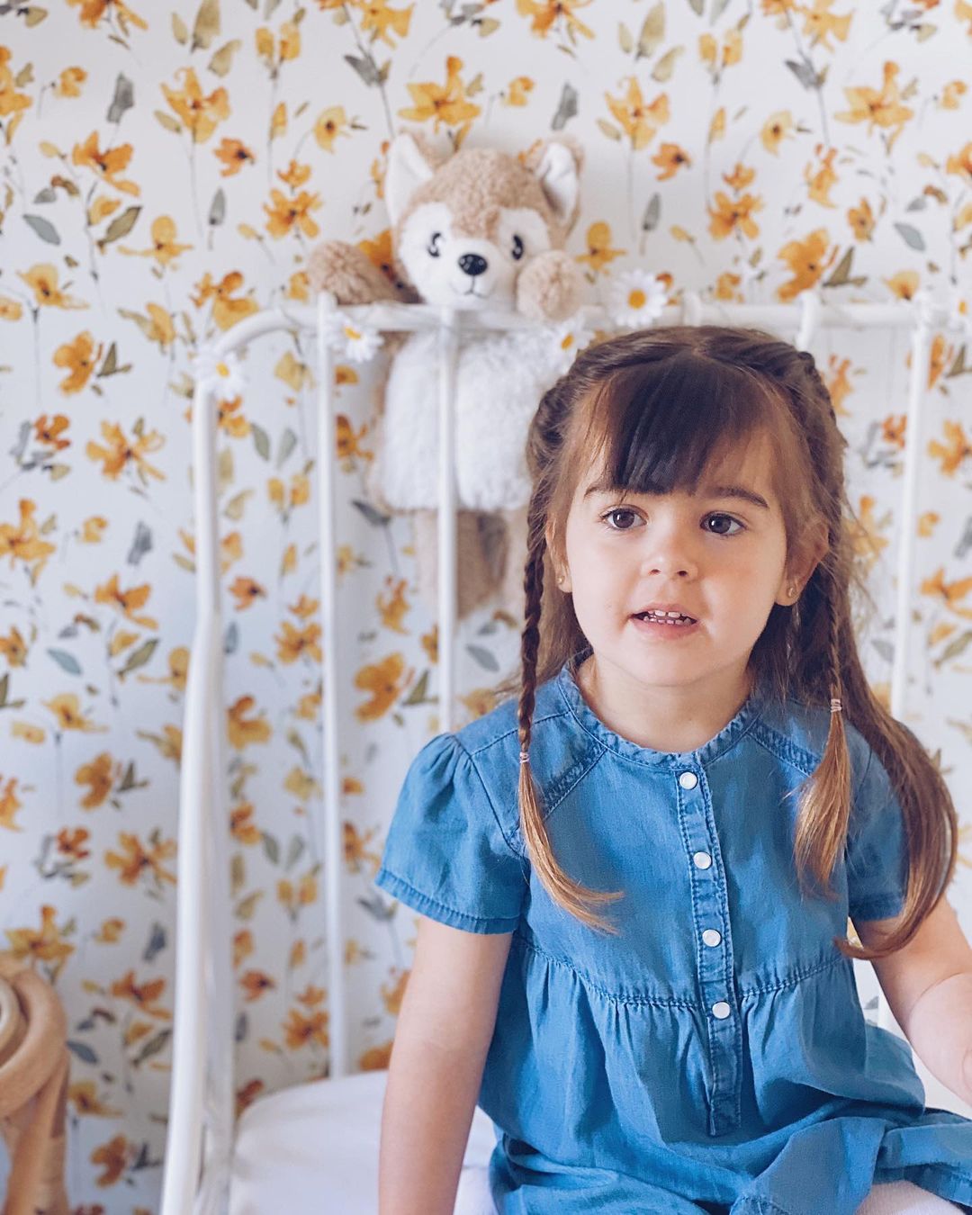 A young girl with pigtails sits in a white crib with floral wallpaper. A stuffed fox toy hangs on the crib.